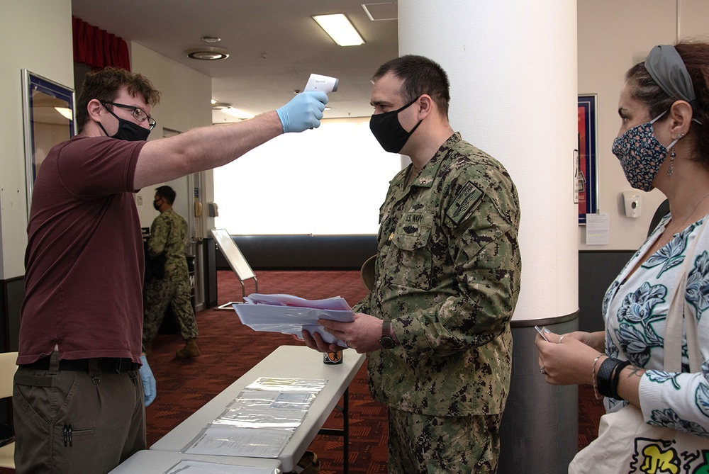 A health screening checkpoint is being conducted, likely during the COVID-19 pandemic. A civilian worker wearing blue latex gloves is using a non-contact infrared thermometer to check the temperature of a U.S. Navy service member in camouflage uniform. A woman in a floral mask waits in line behind the service member.