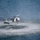 HSC-3 Search and Rescue (SAR) Training A military helicopter, consistent with an MH-60 Seahawk variant, is hovering at very low altitude directly over open water. The rotor wash is creating a large circular disturbance pattern on the water surface, with spray and ripples radiating outward. A crew member appears to be visible in the open side door, suggesting a rescue, insertion, or hoist operation in progress.