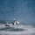 HSC-3 Search and Rescue (SAR) Training A military helicopter, appearing to be an MH-60 Seahawk, hovers at low altitude over open water. A single person is visible standing or suspended beneath the helicopter, surrounded by rotor wash creating circular wave patterns on the water surface. This appears to be a search and rescue or combat rescue swimmer training operation.