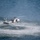HSC-3 Search and Rescue (SAR) Training A military helicopter, consistent with an MH-60 Seahawk variant, hovers at low altitude over open ocean water. The rotor wash is creating a large circular disturbance pattern on the water surface. A person or object appears to be suspended below the helicopter on a hoist line, suggesting a search and rescue or combat rescue operation.