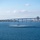 HSC-3 Search and Rescue (SAR) Training A military helicopter hovers at very low altitude over a bay, creating rotor wash disturbance on the water surface. A long concrete bridge spans the background across the bay, with a city skyline visible on the right side. The scene appears to be San Diego Bay with the Coronado Bridge and downtown San Diego skyline.