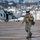 HSC-3 Search and Rescue (SAR) Training A U.S. military aviator in full flight gear walks across a wet flight deck or tarmac, with a gray military helicopter visible in the background. The pilot is wearing an olive green flight suit, tactical vest, flight helmet with visor, and carrying a flight bag. The background shows an urban coastal cityscape, suggesting a naval base or port facility.