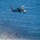 HSC-3 Search and Rescue (SAR) Training A military helicopter, appearing to be an HH-60 Seahawk or similar variant, hovers over ocean surf while conducting a hoist rescue or training operation. A person in dark gear with what appears to be a red element is suspended on a hoist cable below the aircraft near the breaking waves. At least two additional individuals are visible in the water near the shoreline.