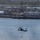 HSC-3 Search and Rescue (SAR) Training A military helicopter, consistent with an MH-60 Seahawk, hovers over a body of water while a person is being lowered or hoisted on a cable below it. The rotor wash is creating a visible circular disturbance pattern on the water surface. The background shows a military or naval installation with industrial buildings, piers, and green parkland beyond.