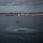 HSC-3 Search and Rescue (SAR) Training A military or rescue helicopter hovers low over a bay or harbor at dusk, with a figure suspended below it on a hoist line. The rotor wash is creating a large circular disturbance pattern on the water surface below. A large bridge and an illuminated urban waterfront skyline are visible in the background under overcast skies.
