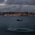 HSC-3 Search and Rescue (SAR) Training A military or rescue helicopter hovers low over a bay at dusk, with a person visibly suspended beneath it on a hoist or rope. A large curved bridge spans the background, with a lit cityscape behind it. The scene appears to depict a training or rescue operation conducted over open water in low-light conditions.