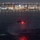 HSC-3 Search and Rescue (SAR) Training A military or law enforcement helicopter flies low over a harbor at night, illuminated by a bright red navigation/warning light. A large cable-stayed or arch bridge spans the background, with a densely lit urban skyline behind it. The helicopter's rotor wash is visibly disturbing the water surface below.