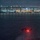 HSC-3 Search and Rescue (SAR) Training A military helicopter, consistent with a UH-60 Black Hawk silhouette, hovers low over a dark bay at night with a figure suspended below on a hoist or rope. A large cable-stayed or arch bridge and an illuminated city skyline are visible in the background, consistent with San Diego Bay and the Coronado Bridge. The scene appears to depict a nighttime hoist or fast-rope training or rescue operation over open water.