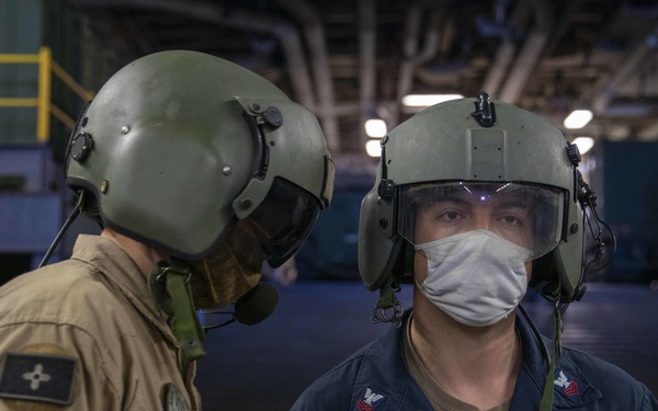 Two military personnel, likely U.S. Navy sailors, are standing in what appears to be a ship's hangar bay. Both are wearing aviation crew helmets with visors; the foreground individual is in a dark blue coverall uniform and wearing a face mask, while the background individual wears a tan/khaki flight suit. The setting suggests an aircraft carrier or amphibious assault ship interior.