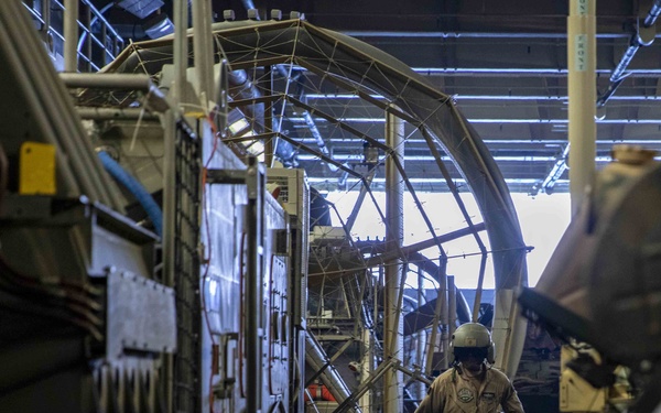 A military or aviation technician in a flight suit and helmet works inside what appears to be a large aircraft hangar or the interior of a military transport aircraft. The environment is industrial with structural framework arches, equipment panels, and various mechanical components visible. The lighting is dim with fluorescent overhead lights illuminating the cavernous space.
