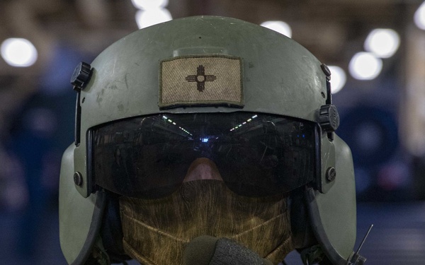 A military personnel member is shown wearing a tactical flight helmet with a dark visor, a face covering, and a boom microphone headset. A tan patch featuring the Zia sun symbol (associated with New Mexico) is visible on the front of the helmet. The individual appears to be standing in a large indoor facility, likely a hangar bay.
