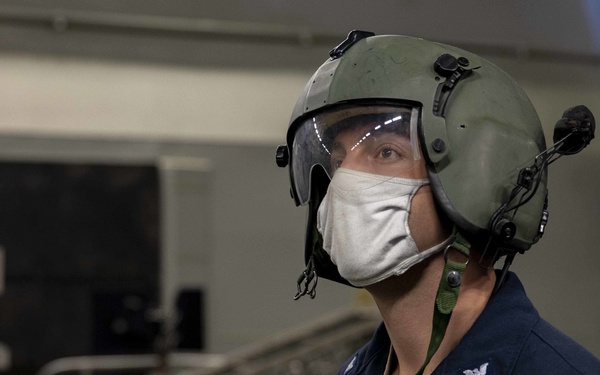 A U.S. Navy sailor wearing a green flight deck helmet with a clear visor and a white cloth face mask is photographed in what appears to be a ship's hangar bay. The sailor's name tag reads 'PETER MAY' with the rating 'ET1' and branch 'USN' visible on their dark blue coveralls. The background shows industrial equipment and structural elements consistent with a naval vessel interior.