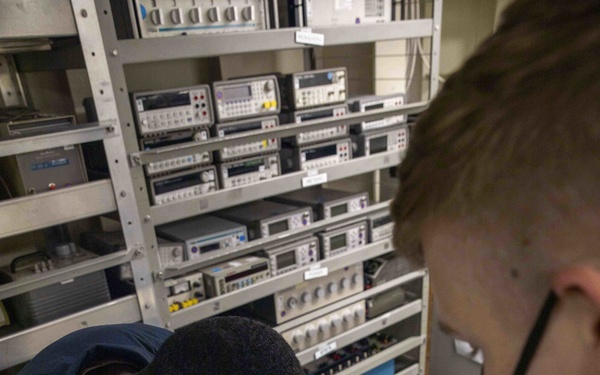 Two individuals wearing dark uniforms and face masks are working in what appears to be a military or technical calibration laboratory. They are reviewing documents in a binder while examining a precision instrument housed in a protective case. The background contains multiple racks of electronic test and measurement equipment.