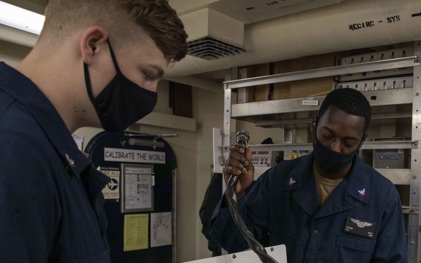 Two U.S. Navy personnel in dark blue coveralls and black face masks are working together in what appears to be an avionics or calibration shop aboard a naval vessel. They are handling a white equipment box with cables and connectors, likely performing maintenance or testing procedures. The workspace contains shelving units with equipment, reference materials, and a sign reading 'CALIBRATE THE WORLD'.