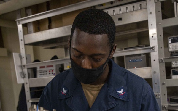 A U.S. Navy Aviation Electronics Technician Third Class (AT3) named Tyrese Holt is working with cables and connectors on what appears to be avionics equipment. He is wearing a dark blue Navy coverall uniform with rank insignia and an aviation warfare device, along with a black face mask. The setting appears to be a technical workspace or shop aboard a naval vessel, surrounded by rack-mounted electronic equipment.