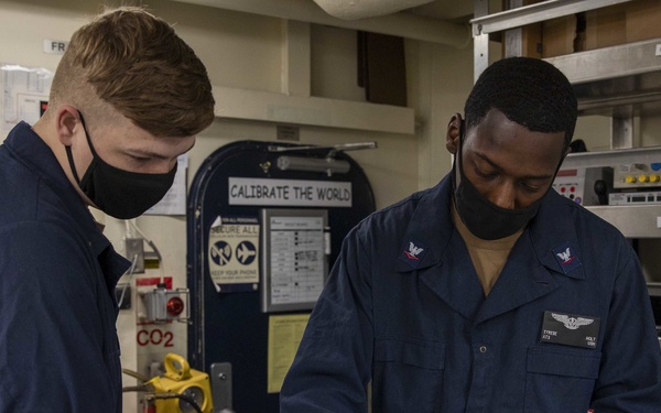 Two U.S. Navy personnel in dark blue coveralls and black face masks are working together on an electronic test or calibration device inside what appears to be a shipboard workspace. The individual on the right, whose name tag reads 'HOLT' with an ATS rating badge, is operating a panel with multiple colored knobs and switches, while the person on the left holds a document or checklist. The workspace contains safety equipment, labeled storage shelves, and a sign reading 'CALIBRATE THE WORLD.'