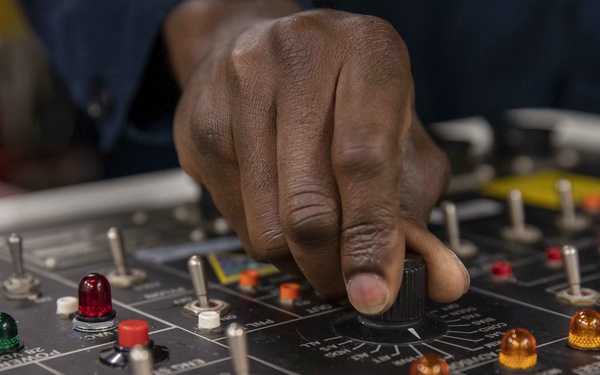 A person's hand is adjusting a knob on a complex control panel or equipment board. The panel features numerous indicator lights in red, orange, green, and amber colors, along with toggle switches and rotary knobs. The equipment appears to be a technical or industrial control system, possibly related to broadcasting, aviation training, or electrical systems.