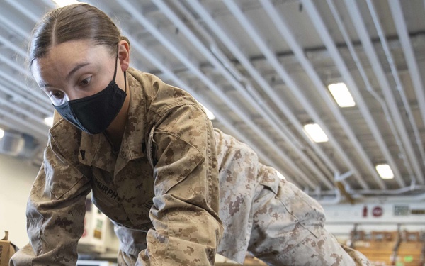 A female Marine in MARPAT desert camouflage uniform and black face mask is kneeling on the floor of what appears to be a ship's hangar bay, handling linked ammunition belts laid out in rows. Her name tape reads 'MURPHY' and she appears to be counting, organizing, or inspecting the ammunition. The setting includes military equipment, ammo cans, and a ship's crest visible on the wall in the background.