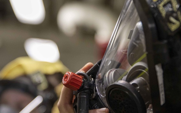 The image shows personnel in firefighting or emergency response gear, wearing full-face respirator masks connected to breathing apparatus hoses. The foreground subject is adjusting or connecting a regulator fitting with a distinctive red knob on their SCBA (Self-Contained Breathing Apparatus) mask. At least two individuals are visible, both equipped with yellow protective gear consistent with firefighting or damage control operations.