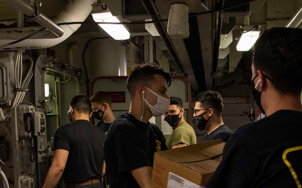 A group of military personnel aboard what appears to be a naval vessel are engaged in a supply chain or logistics operation, passing boxes through a corridor. All individuals are wearing face masks, suggesting COVID-19 era protocols. At least one person is visibly wearing a 'Food Service Attendant' shirt, indicating this is a food or supply resupply operation.