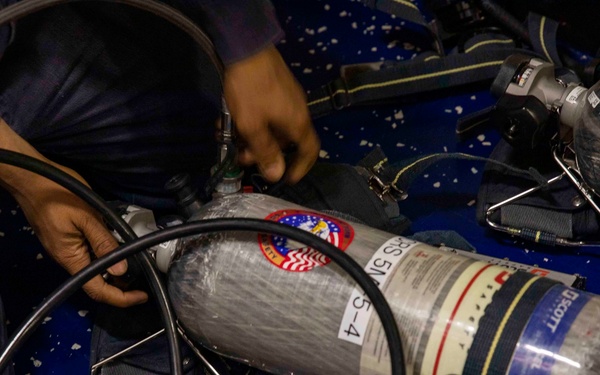 A person in dark blue uniform is handling Self-Contained Breathing Apparatus (SCBA) equipment, likely performing maintenance, inspection, or preparation. Multiple carbon fiber SCBA cylinders branded 'Scott' are visible, laid out on a blue surface. The cylinders bear circular patches featuring an American flag design, suggesting a U.S. military or emergency services context.