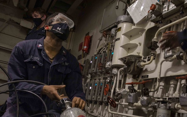 A U.S. Navy sailor wearing a face mask, safety goggles, and a ship's ball cap is working with SCBA (Self-Contained Breathing Apparatus) cylinders in what appears to be a shipboard engineering or damage control space. The sailor, whose name tag reads 'LAPIERRE,' is handling compressed air cylinders marked with Scott branding and identification labels. The setting is a confined mechanical space aboard a naval vessel with extensive piping, valves, and control panels visible on the bulkhead.