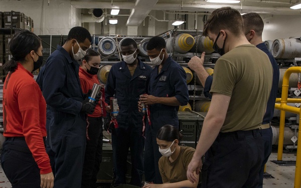 A group of approximately seven military personnel, wearing face masks, are gathered in what appears to be a ship's weapons/ordnance storage compartment. They are handling cylindrical components, likely fuzes or ordnance parts, with one person crouching over a military ammunition crate. The setting contains large munitions or torpedoes stored on racks in the background.