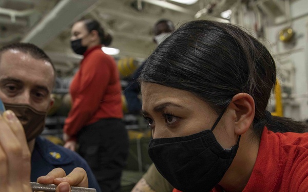 Military personnel wearing red shirts and black face masks are working in what appears to be a ship's hangar bay or weapons handling area. A woman in the foreground is closely examining or working on a cylindrical blue object using a small tool, assisted by a male colleague. Additional personnel in red shirts are visible in the background.