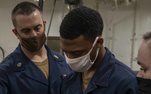 Three U.S. Navy personnel in blue coveralls and face masks are gathered in what appears to be a shipboard workspace or engineering space. Two sailors and one individual in a red shirt are examining or working on a small mechanical component or tool. The scene appears to be a training or maintenance activity aboard a naval vessel.