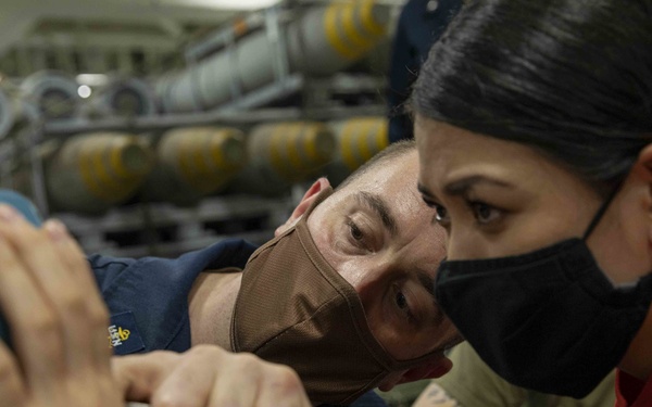 Two military personnel wearing face masks are closely examining or working on a large cylindrical object, likely ordnance or a munition component. Both individuals are wearing navy blue uniforms with visible insignia patches. The setting appears to be a weapons storage or ordnance handling area aboard a naval vessel, with multiple munitions visible in the background.