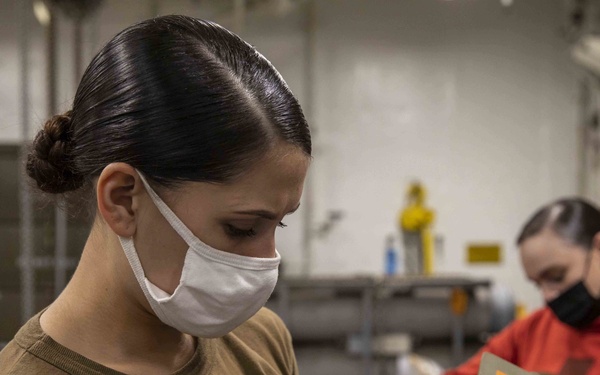 Two individuals wearing face masks are working in what appears to be a military or industrial facility. The foreground person, wearing a tan/olive t-shirt, is examining a small cylindrical metal component alongside an olive-drab military-style box with orange warning labels. A second person in a red hoodie with a black mask works in the background.
