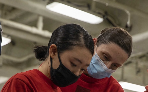 Two individuals wearing matching red long-sleeve shirts and face masks are examining documents together in what appears to be an industrial or military interior space. The person in the background, whose shirt displays chevron rank insignia consistent with a U.S. Navy Petty Officer, appears to be supervising or instructing the person in the foreground. They are referencing a stack of papers near a large cylindrical mechanical component.