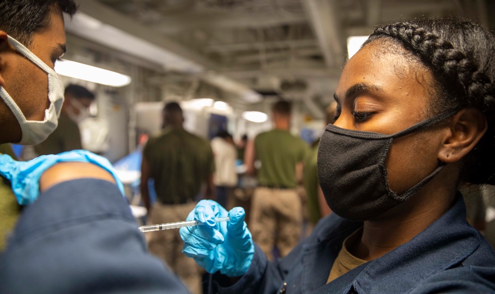 A medical professional wearing blue latex gloves and a white surgical mask is administering an injection to a service member wearing a black face mask and dark blue uniform. The setting appears to be a military vessel or installation, with additional personnel visible in the background. The scene is consistent with a military vaccination or medical treatment event.