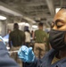 A medical professional wearing blue latex gloves and a white surgical mask is administering an injection to a service member wearing a black face mask and dark blue uniform. The setting appears to be a military vessel or installation, with additional personnel visible in the background. The scene is consistent with a military vaccination or medical treatment event.
