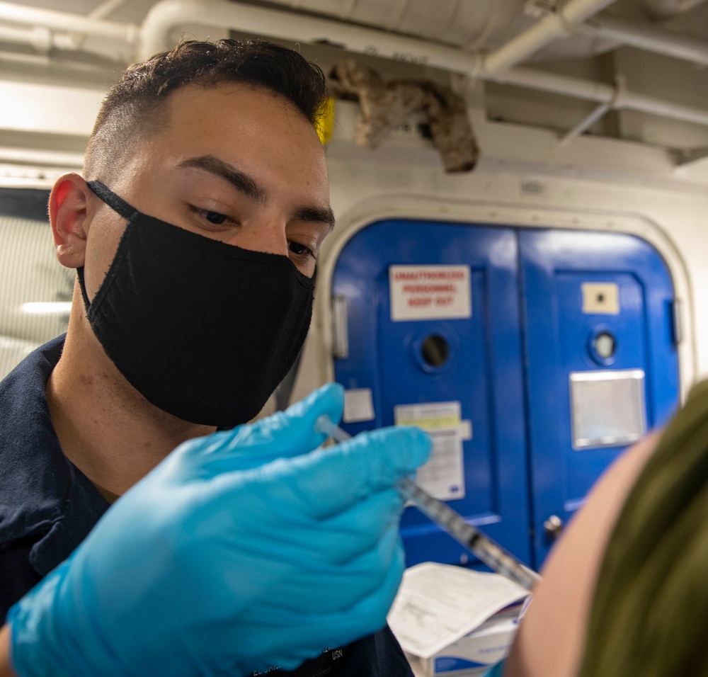 A medical personnel wearing a black face mask and blue nitrile gloves is administering an injection to a patient in what appears to be a military naval vessel setting. The blue watertight door with a 'Unauthorized Personnel Keep Out' sign in the background is characteristic of a U.S. Navy ship's interior. The scene depicts a vaccination or medical procedure being conducted aboard a naval vessel.