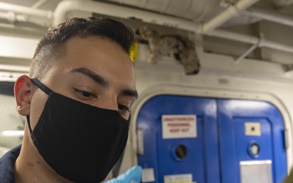A medical personnel wearing a black face mask and blue nitrile gloves is administering an injection to a patient in what appears to be a military naval vessel setting. The blue watertight door with a 'Unauthorized Personnel Keep Out' sign in the background is characteristic of a U.S. Navy ship's interior. The scene depicts a vaccination or medical procedure being conducted aboard a naval vessel.