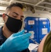 A medical personnel wearing a black face mask and blue nitrile gloves is administering an injection to a patient in what appears to be a military naval vessel setting. The blue watertight door with a 'Unauthorized Personnel Keep Out' sign in the background is characteristic of a U.S. Navy ship's interior. The scene depicts a vaccination or medical procedure being conducted aboard a naval vessel.