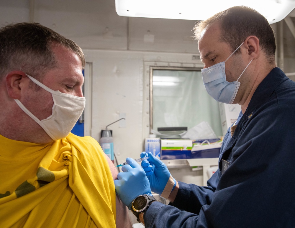 A medical professional in a dark navy uniform is administering an injection to a patient wearing a yellow shirt in what appears to be a medical facility or sick bay. Both individuals are wearing face masks, and the administrator is wearing blue nitrile gloves. The setting appears to be a military or naval medical environment based on the institutional interior and uniform.