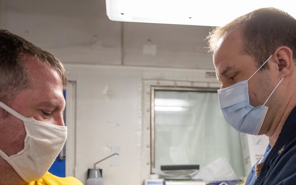 A medical professional in a dark navy uniform is administering an injection to a patient wearing a yellow shirt in what appears to be a medical facility or sick bay. Both individuals are wearing face masks, and the administrator is wearing blue nitrile gloves. The setting appears to be a military or naval medical environment based on the institutional interior and uniform.