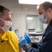 A medical professional in a dark navy uniform is administering an injection to a patient wearing a yellow shirt in what appears to be a medical facility or sick bay. Both individuals are wearing face masks, and the administrator is wearing blue nitrile gloves. The setting appears to be a military or naval medical environment based on the institutional interior and uniform.