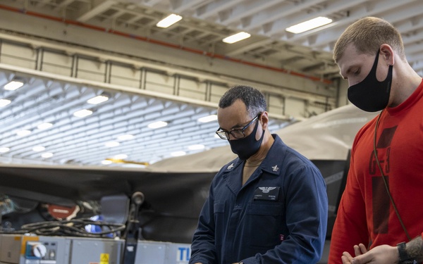 Two military personnel wearing face masks are working at a table in what appears to be an aircraft carrier hangar bay. They are handling green military ammunition cans and related equipment, with at least one stealth aircraft (appearing to be an F-35) visible in the background. The setting suggests an ordnance or equipment preparation activity aboard a naval vessel.