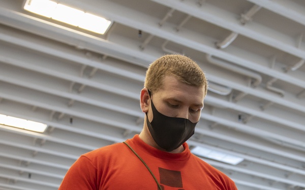A male individual wearing a red long-sleeve shirt marked 'O-3' and a black face mask is working in what appears to be a military hangar bay, likely aboard a naval vessel. He is handling a cord or lanyard over what appears to be a military-green ammunition or equipment case. The environment features fluorescent lighting, overhead piping, and aircraft visible in the background.