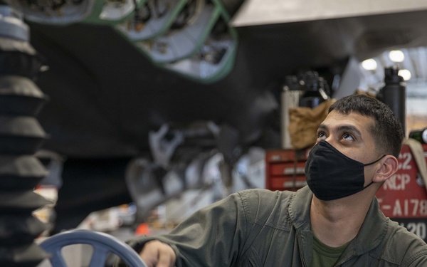 A U.S. Marine Corps Staff Sergeant named Hernandez, wearing a black face mask and olive green flight suit, is performing maintenance work underneath a military aircraft in a hangar. He is looking upward at the aircraft's undercarriage while resting his hand on a blue wheel-shaped ground support equipment piece. The setting appears to be an active maintenance bay with tools and equipment visible in the background.