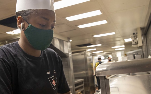 A kitchen worker wearing a white chef's hat, green face mask, and black t-shirt is preparing raw meat on a cutting surface in what appears to be a large institutional or military galley kitchen. The worker also has a wireless earbud in their ear. The background shows a long stainless steel kitchen corridor with additional staff working in the distance.
