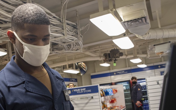A U.S. Navy sailor in blue coveralls and a white face mask operates a point-of-sale terminal at what appears to be a ship's store (Navy Exchange). The signage in the background reads 'YOUR SHIP YOUR STORE,' confirming this is an onboard retail facility on a naval vessel. A second masked individual is visible in the background near refrigerated drink displays.