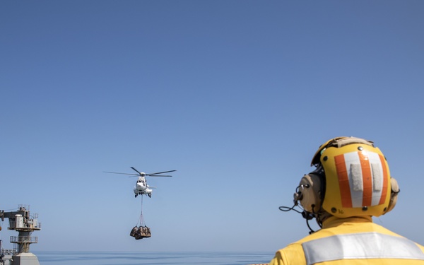 A U.S. Navy Flight Deck Chief Petty Officer (CPO) in yellow safety gear observes a vertical replenishment (VERTREP) operation from the deck of what appears to be an aircraft carrier. A helicopter is airlifting a cargo load between the carrier and an alongside replenishment vessel during an underway replenishment (UNREP) at sea. The operation is taking place in calm, clear blue-sky conditions on open ocean.