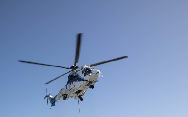 A flight deck crew member aboard a naval vessel (likely an LHD-class amphibious assault ship) guides a civilian contract helicopter during a vertical replenishment (VERTREP) operation. The helicopter, marked '07' and painted white and blue, is airlifting cargo nets filled with supplies to the flight deck. Multiple netted cargo pallets are already staged on the deck awaiting distribution.