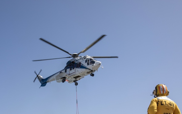 A helicopter is conducting a vertical replenishment (VERTREP) operation over what appears to be a naval vessel's deck, lowering a cargo net filled with supplies. Two personnel are visible on deck — one in a yellow vest marked 'FLT DV CPO' and another in green utilities with a white helmet — overseeing the cargo transfer. Multiple netted cargo pallets are stacked on the deck to the right, indicating an active logistics resupply operation.