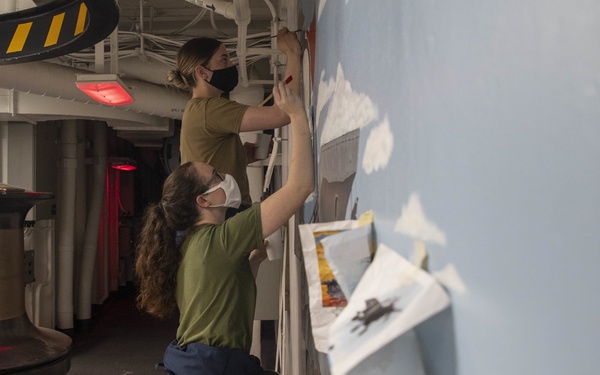 Two individuals wearing face masks are painting a large mural on a wall inside what appears to be a naval vessel or industrial corridor. One person stands on a chair to reach the upper portion of the mural, while the other works on the lower section. Reference photographs are taped to the wall to guide their work, and the mural appears to depict an arctic or winter landscape scene.
