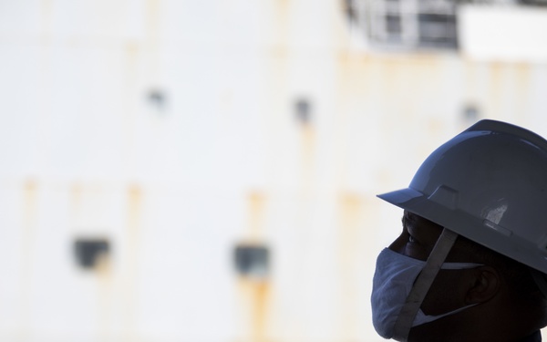 A worker is shown in profile view wearing personal protective equipment including a hard hat and face mask. They appear to be stationed near a large industrial structure, likely a ship or storage tank, which shows visible rust staining on its white surface. The image is dramatically lit with the subject partially silhouetted against the bright background.
